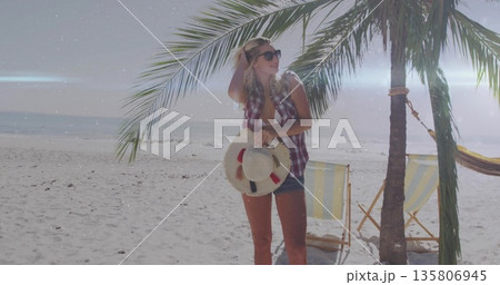 Holding straw hat and touching hair, woman wearing sunglasses posing on sandy beach by hammock 135806945