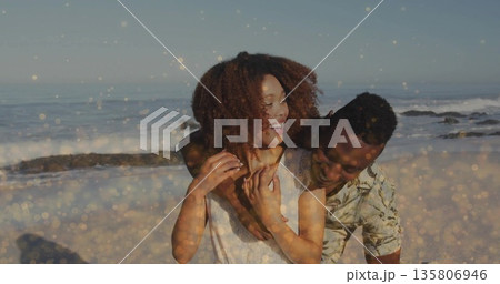 Hugging laughing couple wearing sleeveless top and tropical shirt at sandy beach, sparkling waves 135806946