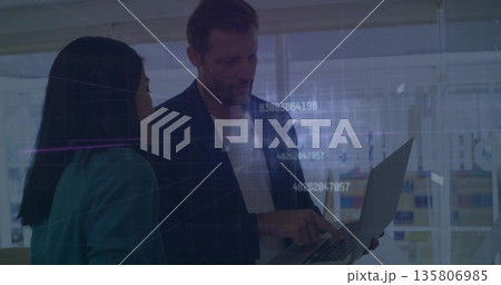 Pointing man and woman wearing suits collaborating on laptop in modern office, with data overlay 135806985