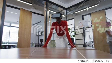 Leaning woman wearing VR headset placing hands on table in glass meeting room, with sticky notes 135807045