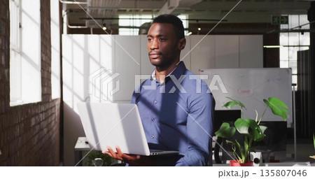 Standing office worker holding silver laptop in open-plan office, with whiteboard and plant Standing office worker holding silver laptop in open-plan office, with whiteboard and plant 135807046