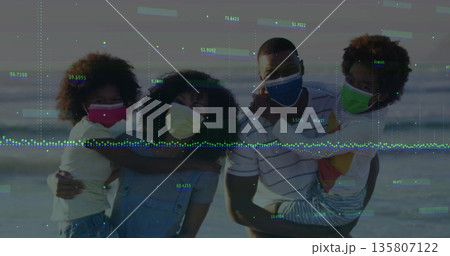 Embracing family wearing colorful face coverings standing on sandy beach at dusk, ocean backdrop 135807122