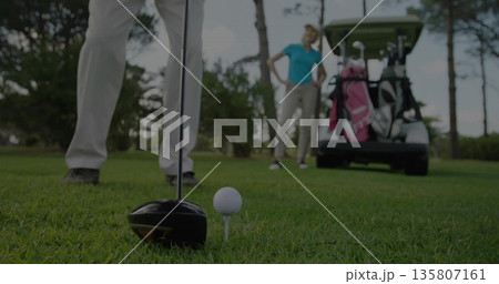 Positioning driver club head behind golf ball on tee at tee box, with golf cart, bags 135807161