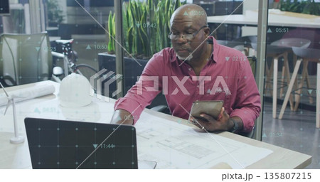 Engineer in red shirt operating tablet and laptop in office, with blueprints and digital overlays Engineer in red shirt operating tablet and laptop in office, with blueprints and digital overlays 135807215