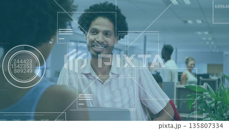Smiling man leaning on desk talking at open-plan office with laptop, digital overlay graphics Smiling man leaning on desk talking at open-plan office with laptop, digital overlay graphics 135807334
