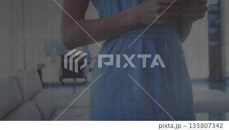 Standing woman in blue dress holding smartphone in lounge area, with sofa, table, chairs and lamp 135807342