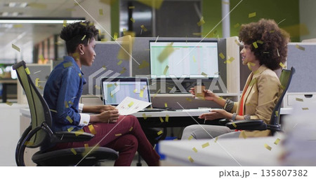 Throwing gold confetti two women in business attire discussing work at office desk, with laptop 135807382
