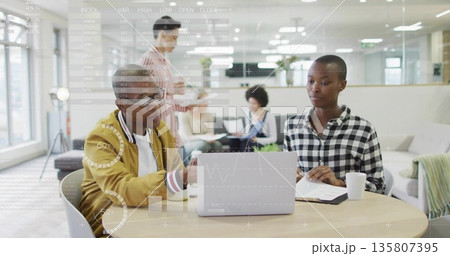 Collaborating pair in casual attire working on laptop and documents at office table, with charts Collaborating pair in casual attire working on laptop and documents at office table, with charts 135807395