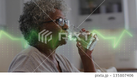 Lifting senior woman in glasses raising clear glass to lips at kitchen, with green heartbeat line 135807406