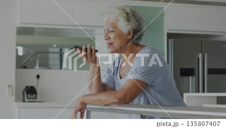 Leaning senior woman wearing blue top holding smartphone near mouth in modern kitchen, with railing Leaning senior woman wearing blue top holding smartphone near mouth in modern kitchen, with railing 135807407
