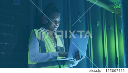 Technician checking racks in data center, with laptop earmuffs safety vest, copy space 135807563