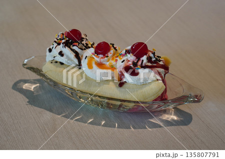 Three flavors of ice cream, decorated with whipped cream and cherries and top with orange jam or strawberry jam and slice banana split, served in a boat-shaped glass on wooden floor background, Three flavors of ice cream, decorated with whipped cream and cherries and top with orange jam or strawberry jam and slice banana split, served in a boat-shaped glass on wooden floor background, 135807791