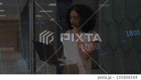 Holding laptop, woman in blouse analyzing chart overlay with gridlines in office, glass partitions Holding laptop, woman in blouse analyzing chart overlay with gridlines in office, glass partitions 135808584