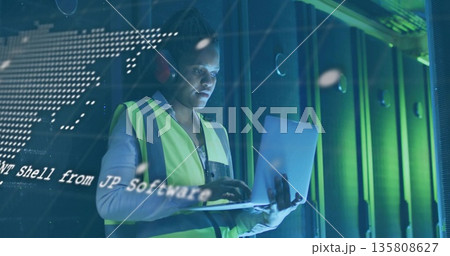 Tech wearing hi-vis vest, red earmuffs reading laptop code overlay in data center before racks Tech wearing hi-vis vest, red earmuffs reading laptop code overlay in data center before racks 135808627