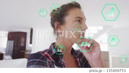 Paint-smeared woman in plaid shirt holding chin in interior with floating green icons, copy space Paint-smeared woman in plaid shirt holding chin in interior with floating green icons, copy space 135809753
