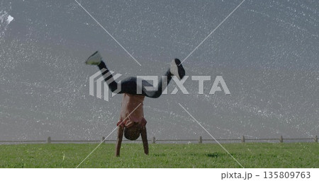 Performing handstand boy in pink t-shirt balancing on hands on grassy field, with wooden fence 135809763