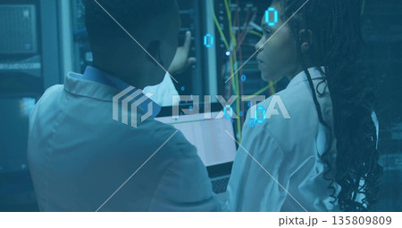 Two IT techs wearing lab coats inspecting server racks in data center, with network cables, laptop Two IT techs wearing lab coats inspecting server racks in data center, with network cables, laptop 135809809