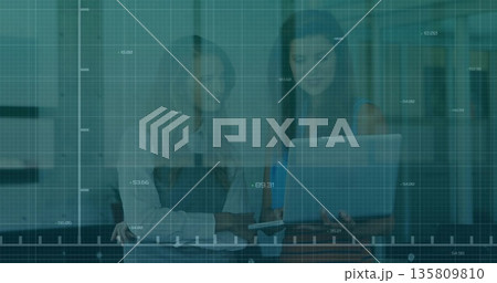 Two women in business attire analyzing grid overlay on desk in office, with laptop and smartphone Two women in business attire analyzing grid overlay on desk in office, with laptop and smartphone 135809810