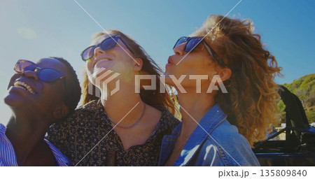 Leaning trio of friends in sunglasses tilting heads back in convertible by seaside road, sun flare Leaning trio of friends in sunglasses tilting heads back in convertible by seaside road, sun flare 135809840