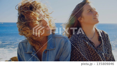 Standing women gazing toward sun on rocky shore, with denim jacket, patterned blouse, necklace Standing women gazing toward sun on rocky shore, with denim jacket, patterned blouse, necklace 135809846