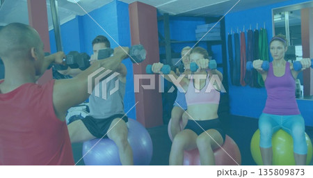 Performing front dumbbell raises group wearing sportswear sitting on stability balls in gym studio Performing front dumbbell raises group wearing sportswear sitting on stability balls in gym studio 135809873