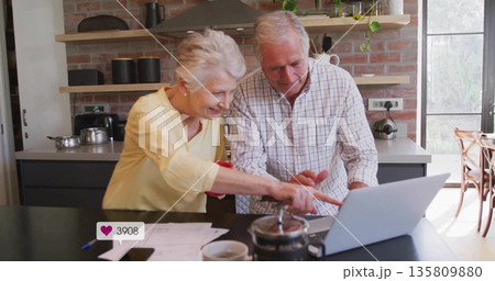 Leaning senior couple working on laptop at home kitchen island with papers pen French press mug 135809880