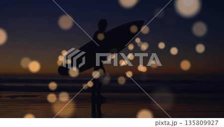 Walking wetsuit-clad surfer holding surfboard on wet sand at dusk, with bokeh orbs reflecting sky Walking wetsuit-clad surfer holding surfboard on wet sand at dusk, with bokeh orbs reflecting sky 135809927