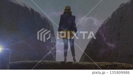Standing explorer wearing wide-brimmed hat and dark jacket on rocky ledge at night, with starry sky Standing explorer wearing wide-brimmed hat and dark jacket on rocky ledge at night, with starry sky 135809986