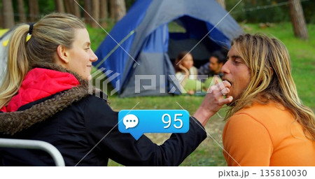 Feeding camper wearing red-hooded coat giving snack in pine clearing, with blue tent and green mat Feeding camper wearing red-hooded coat giving snack in pine clearing, with blue tent and green mat 135810030