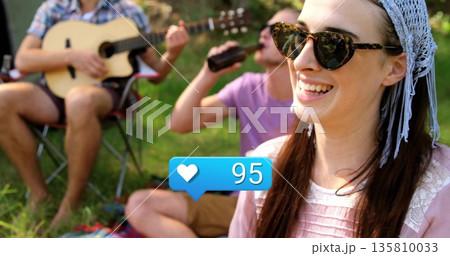 Smiling woman wearing sunglasses, headscarf sitting in grassy clearing, with guitar, copy space 135810033