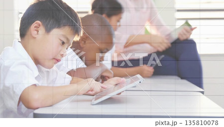 Tapping Asian boy focusing on tablet at white desk in classroom, with horizontal blinds, copy space 135810078