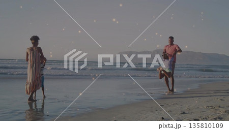 Family walking on beach at sunset with mother wearing dress holding son, father swinging daughter 135810109