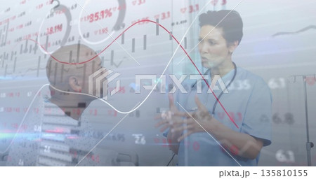 Gesturing Asian doctor in scrubs explaining financial charts in exam room, stethoscope, IV stand 135810155