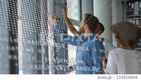 Collaborating professionals sketching diagrams on glass whiteboard in office with sticky notes 135810698