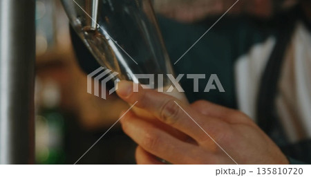 Pouring bartender hands filling tall beer glass at bar interior, with beer tap and frothy head Pouring bartender hands filling tall beer glass at bar interior, with beer tap and frothy head 135810720