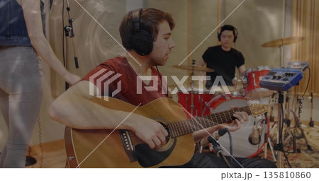 Strumming guitarist in red tee and headphones in studio, with acoustic guitar and drum kit Strumming guitarist in red tee and headphones in studio, with acoustic guitar and drum kit 135810860