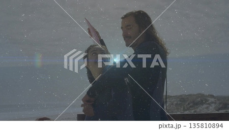 Hugging couple in black wetsuits standing on wooden deck over choppy ocean, holding pink surfboard Hugging couple in black wetsuits standing on wooden deck over choppy ocean, holding pink surfboard 135810894