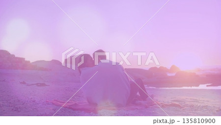 Sitting couple sharing light-colored blanket on pink fringed beach blanket at sunset, pastel light Sitting couple sharing light-colored blanket on pink fringed beach blanket at sunset, pastel light 135810900