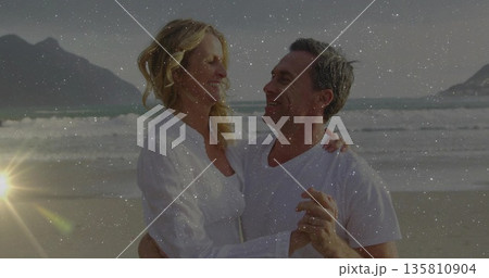 Smiling senior couple embracing near water's edge on sandy beach during sunset, with wedding rings Smiling senior couple embracing near water's edge on sandy beach during sunset, with wedding rings 135810904