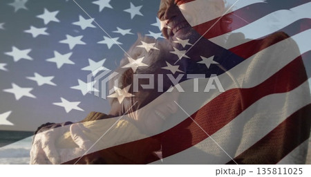 Hugging senior couple wearing knit sweaters on beach, with US flag overlay, rolling ocean waves Hugging senior couple wearing knit sweaters on beach, with US flag overlay, rolling ocean waves 135811025