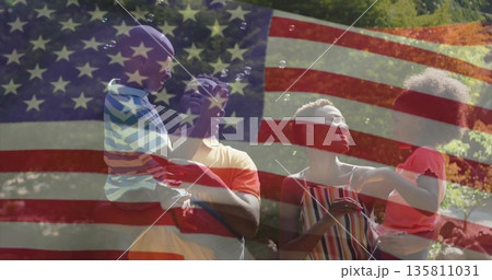 Family of four blowing bubbles with wands in sunlit backyard under faded American flag overlay 135811031