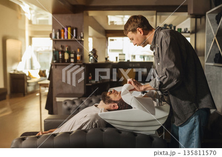 Barber washing client hair with shampoo at salon sink. Professional male grooming service, relaxing head wash process, close up of hairstyling care in modern barbershop. Barber washing client hair with shampoo at salon sink. Professional male grooming service, relaxing head wash process, close up of hairstyling care in modern barbershop. 135811570