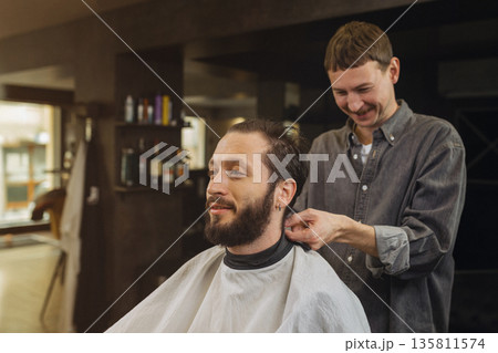 Barber preparing male client for haircut in modern barbershop. Professional barber service, beard grooming, men hairstyle care and lifestyle concept. 135811574