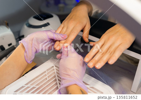 Close up of professional manicure process. Nail technician wearing gloves holding female hands with pink gel polish in beauty salon. Nail care, hygiene, spa treatment concept. Close up of professional manicure process. Nail technician wearing gloves holding female hands with pink gel polish in beauty salon. Nail care, hygiene, spa treatment concept. 135811652