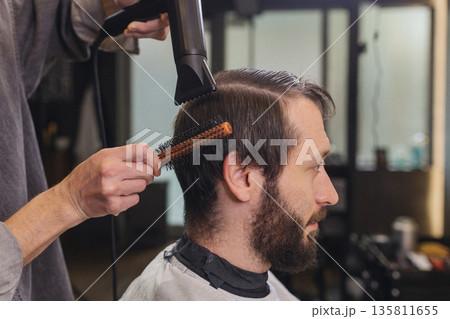 Professional barber styling male hair with hair dryer and brush in modern barbershop. Men grooming service, haircut process, beard care and lifestyle concept. Professional barber styling male hair with hair dryer and brush in modern barbershop. Men grooming service, haircut process, beard care and lifestyle concept. 135811655