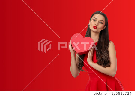 Woman in red dress holds heart-shaped gift against red backdrop Woman in red dress holds heart-shaped gift against red backdrop 135811724