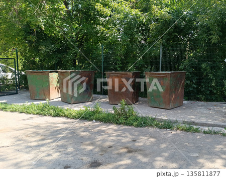 Row of heavily rusted industrial trash bins sits in the bright sun, surrounded by lush green trees and a chain-link fence. Waste disposal 135811877