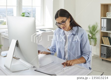 Female accountant looking though business documents at office desk with computer 135813214