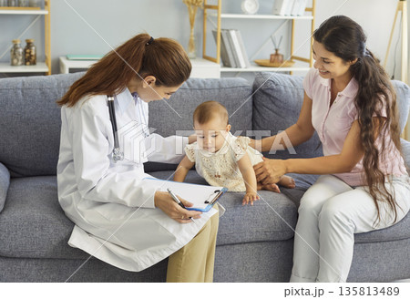Home visit pediatric checkup doctor takes notes as mother supports curious baby 135813489