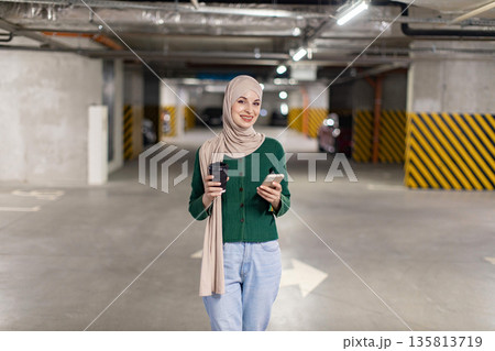 A smiling woman in a hijab holds a coffee cup and smartphone in a parking garage A smiling woman in a hijab holds a coffee cup and smartphone in a parking garage 135813719